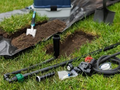 A collection of tools and components for installing an automatic lawn watering system, including hoses, nozzles, and a shovel on a grassy area.